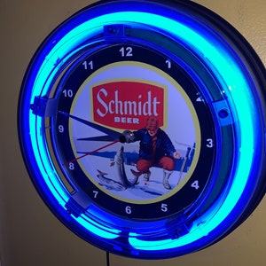 May include: A round wall clock with a black frame and a blue neon light. The clock face features the Schmidt Beer logo and an illustration of a person ice fishing. The clock hands are black, and the numbers are white.