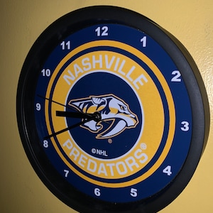 May include: Round wall clock featuring the Nashville Predators logo. The clock face is navy blue and gold, with the team's logo in the center. Black hands and a black frame complete the design. The clock displays the numbers 1 through 12.