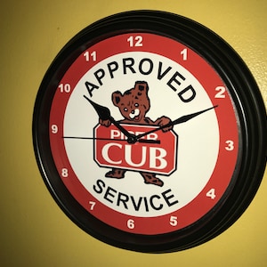 Puede incluir: Reloj de pared redondo con marco negro y esfera roja y blanca. El reloj muestra las palabras "APPROVED CUB SERVICE" y un oso de dibujos animados sosteniendo un letrero. Las manecillas son negras.