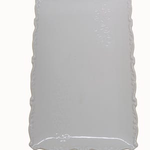 Puede incluir: Una fuente rectangular de cerámica blanca con un borde festoneado decorativo. La fuente presenta detalles florales en relieve y mide aproximadamente 28 cm de alto y 16 cm de ancho. Se ve la marca "L'Infinito".