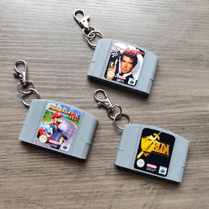 May include: Three grey Nintendo 64 game cartridge keyrings. One features Mario Kart, another has a James Bond image, and the third displays a Zelda graphic. Each keyring has a silver clasp and ring.