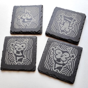 May include: Four square black slate coasters with etched cartoon character designs. Each coaster features a different character with a wavy, circular background. The designs are in a light grey colour, contrasting against the dark slate.