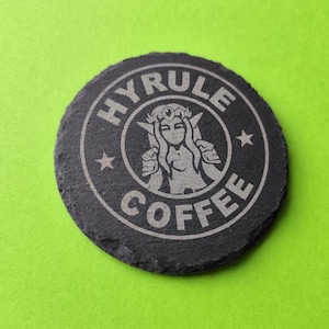 May include: A circular, dark grey slate coaster with the words "HYRULE COFFEE" in a circular design. The centre features a stylized image of a woman. Two small stars are on either side of the image. The coaster is on a bright green background.