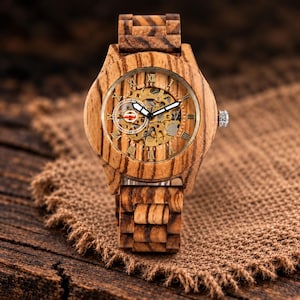 May include: A wristwatch crafted from light and dark brown striped wood featuring a round case and link bracelet. The dial displays gold-toned Roman numerals, white-tipped hands, and an exposed mechanical movement with a small sub-dial.