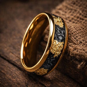 May include: A gold-toned ring featuring a wide band with a polished gold interior and edges. The outer surface displays a textured inlay of black material interspersed with irregular, jagged gold-coloured flakes that create a high-contrast appearance.