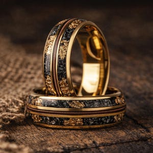Könnte beinhalten: Zwei goldene Eheringe mit schwarzem und goldenem strukturiertem Inlay. Die Ringe haben ein goldenes Band mit einem dünnen kupferfarbenen Streifen und goldenen Akzenten. Die Ringe werden auf einer Holzoberfläche präsentiert.