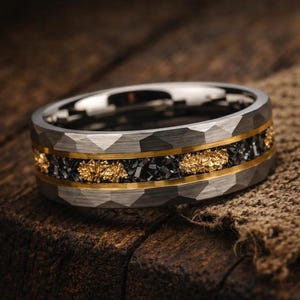 May include: A silver-coloured ring with a textured, faceted band. The ring features a black inlay with gold-coloured accents and a gold-coloured border. The ring is displayed on a wooden surface.