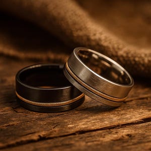 May include: Two wedding rings, one black and one silver, are displayed on a wooden surface. Each ring has a gold-coloured string inlay. The rings are close together, highlighting their contrasting colours and textures.