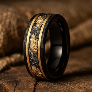 May include: Black ring with gold detailing and a textured central band. The ring has a faceted outer band and gold-coloured inlays. The gold inlays have a rough, natural appearance, adding a unique touch to the jewellery.