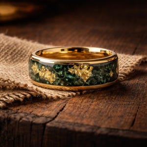 May include: A band ring featuring a polished gold-tone metal interior and edges. The outer surface displays a dark green, textured stone inlay interspersed with irregular, shimmering gold-colored flecks throughout the circumference.