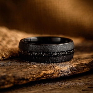 Könnte beinhalten: Ein mattschwarzer Ring mit einer texturierten Oberfläche und einer zentralen horizontalen Einlage aus dunklen, fragmentierten, metallisch wirkenden Partikeln, die ein raues, unebenes Erscheinungsbild um den gesamten Ringumfang erzeugen.