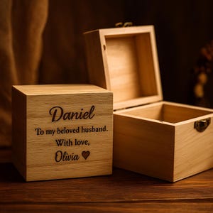 May include: Two light-colored wooden boxes, one closed and one open. The closed box features the engraved text: Daniel, To my beloved husband. With love, Olivia, followed by a small heart symbol. Both boxes have a square shape and a metal latch.