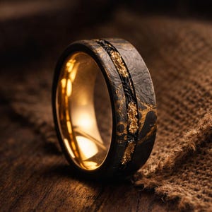 Puede incluir: Primer plano de un anillo negro con detalles dorados. El anillo tiene una superficie texturizada con un interior dorado. Una incrustación dorada recorre el centro del anillo, con una línea negra trenzada a cada lado.