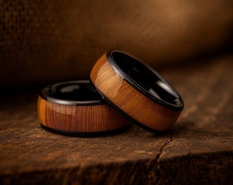 May include: Two rings featuring a polished wood grain exterior inlay set within a smooth black metal band. The rings have a rounded profile and a glossy finish, showcasing the natural vertical lines of the wood grain against the dark metal edges.
