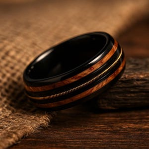 May include: A black ring with a wood grain and a gold-colored string inlay. The ring has a polished finish and a unique design. The ring is a stylish accessory.