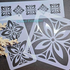 May include: Several white stencils with floral designs. The stencils feature various sizes and patterns of stylized flowers, with a repeating motif. The stencils are arranged on a dark surface, with a piece of coral in the foreground.