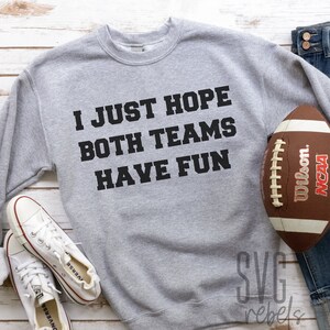 May include: A gray sweatshirt with the text "I JUST HOPE BOTH TEAMS HAVE FUN" printed on the front. A brown football and a pair of blue jeans are also in the image.