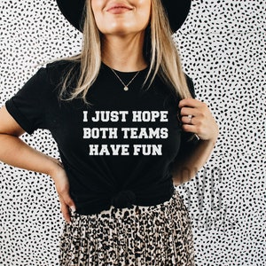 May include: A black t-shirt with white text that reads "I just hope both teams have fun". The person wearing the shirt is standing in front of a white background with black polka dots.