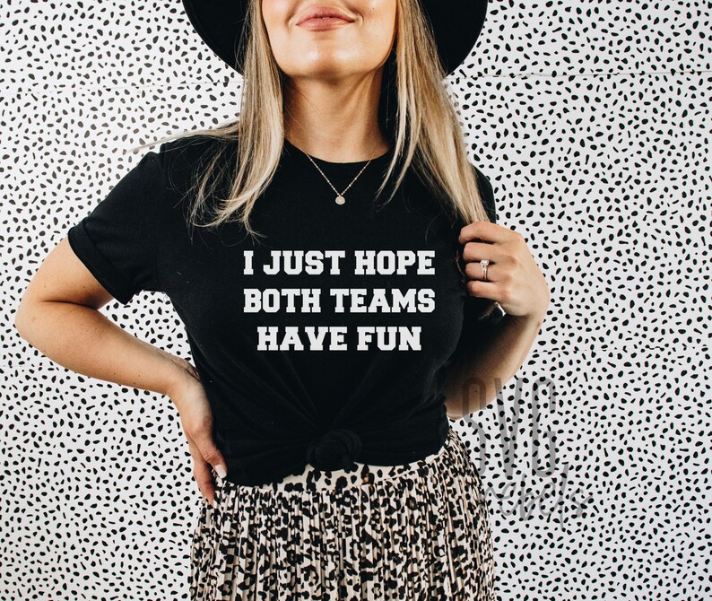 May include: A black t-shirt with white text that reads "I just hope both teams have fun". The person wearing the shirt is standing in front of a white background with black polka dots.