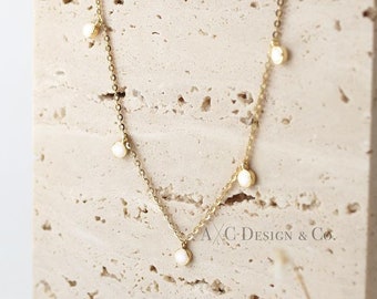 May include: A gold chain necklace with small white round stones hanging from it. The necklace is displayed on a beige textured surface.