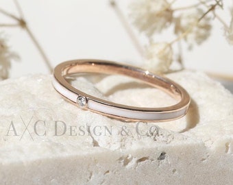 May include: A thin rose gold band ring with a white enamel stripe and a small diamond accent. The ring is sitting on a white marble surface.