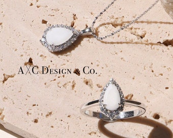 May include: A white teardrop-shaped pendant necklace and a matching ring with a white stone set in a silver band. The jewelry is displayed on a light brown textured surface. The text "AXC Design & Co." is visible on the surface.