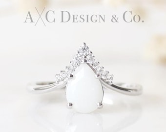 May include: A silver ring with a pear-shaped white opal stone and a V-shaped band of small clear stones.  The ring is on a white surface.  AXC Design & Co.
