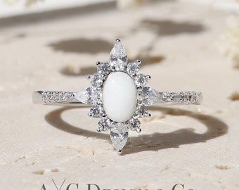 May include: A silver ring with a white oval gemstone surrounded by smaller white gemstones. The ring is on a light brown textured surface. AXC Design & Co.