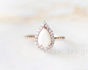 May include: A pearlescent white teardrop-shaped ring with a halo of clear cubic zirconia stones set in a rose gold band.