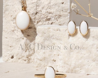May include: A set of white oval gemstone jewelry on a white stone surface. The set includes a pendant necklace, a pair of earrings, and a ring. The jewelry is displayed against a white background with dried white flowers.