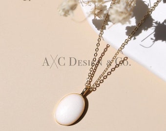 May include: A gold chain necklace with a white oval pendant. The necklace is on a white and beige surface with dried white flowers.
