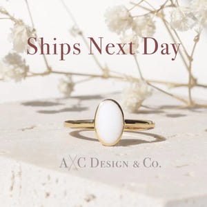 May include: A gold ring with an oval, white stone. The ring is set against a neutral background with the text "Ships Next Day" and "A X C Design & Co."