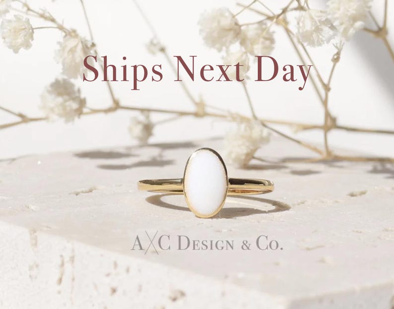 May include: A gold ring with an oval, white stone. The ring is set against a neutral background with the text "Ships Next Day" and "A X C Design & Co."