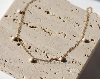 May include: A gold chain necklace with five white round beads hanging from it. The necklace is on a light brown textured surface.