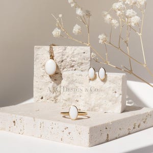 May include: A jewelry set featuring a gold necklace, ring, and earrings with white oval-shaped stones. The jewelry is displayed on a textured, light-colored surface with a sprig of white flowers. The brand name "A&C Design & Co." is visible.