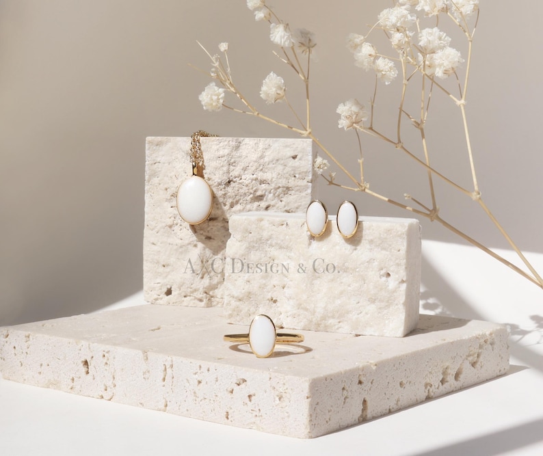 May include: A jewelry set featuring a gold necklace, ring, and earrings with white oval-shaped stones. The jewelry is displayed on a textured, light-colored surface with a sprig of white flowers. The brand name "A&C Design & Co." is visible.