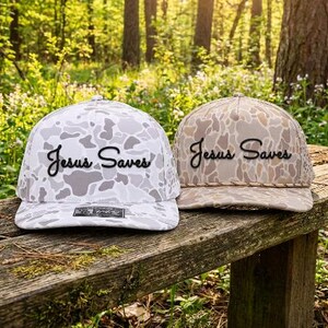 Puede incluir: Dos gorras de b&eacute;isbol con estampado de camuflaje y el texto "Jesus Saves" en letras cursivas negras. La gorra de la izquierda es blanca y gris, mientras que la de la derecha es beige y marr&oacute;n con un detalle de cord&oacute;n trenzado en la base.