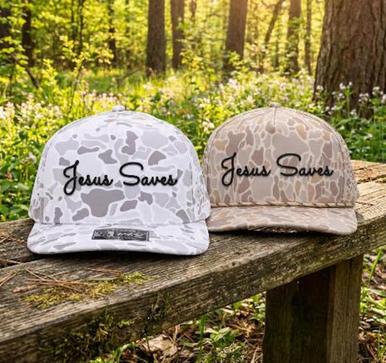 Puede incluir: Dos gorras de b&eacute;isbol con estampado de camuflaje y el texto "Jesus Saves" en letras cursivas negras. La gorra de la izquierda es blanca y gris, mientras que la de la derecha es beige y marr&oacute;n con un detalle de cord&oacute;n trenzado en la base.