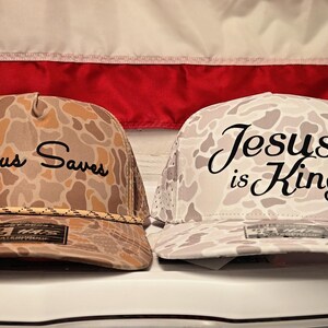 Puede incluir: Dos gorras tipo camionero con patrones de camuflaje. La gorra izquierda es de color canela y marr&oacute;n con el texto negro 'Jesus Saves' y un detalle de cuerda. La gorra derecha es de color blanco y gris con el texto negro 'Jesus is King'.