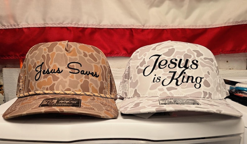 Puede incluir: Dos gorras tipo camionero con patrones de camuflaje. La gorra izquierda es de color canela y marr&oacute;n con el texto negro 'Jesus Saves' y un detalle de cuerda. La gorra derecha es de color blanco y gris con el texto negro 'Jesus is King'.