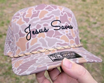 以下が含まれることがあります： ピンク、タン、ホワイトの迷彩柄に黒の刺繍でJesus Savesと書かれた野球帽。帽子のつばにはベージュのロープのディテールがあり、10 10 HATS EST 2023と書かれた黒いステッカーが貼られています。