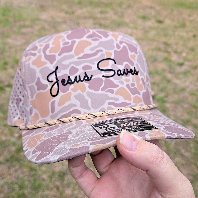 Puede incluir: Una gorra de b&eacute;isbol con un patr&oacute;n de camuflaje en tonos malva, beige y marr&oacute;n claro. El panel frontal muestra el texto 'Jesus Saves' bordado en negro con una fuente cursiva. Un cord&oacute;n fino y moteado se sit&uacute;a sobre la visera de la gorra.