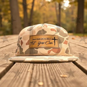May include: A camouflage-patterned baseball cap with a flat brim. The cap features a rectangular patch with the text "GIVE YOUR LIFE TO JESUS 'Til You Can" and a cross symbol. The cap is set against a blurred outdoor background.