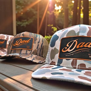 Puede incluir: Tres gorras de béisbol de camuflaje con la palabra "Dad" en un parche de cuero. Las gorras tienen patrones de camuflaje en tonos marrones, verdes y azules. Una gorra tiene el nombre "Wrenleigh Yvonne Searson" y la fecha "DEC 7".