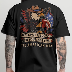 May include: Black t-shirt featuring a patriotic design. The graphic includes an eagle wearing a cowboy hat, holding a drink and an American flag. Text on the shirt reads "WE'LL PUT A BOOT IN YOUR ASS. IT'S THE AMERICAN WAY."