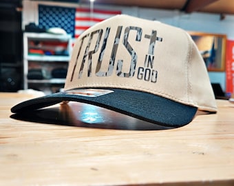 May include: A tan baseball cap with a black brim. The cap features the words "TRUST IN GOD" in a camouflage pattern across the front. The cap is sitting on a wooden surface.