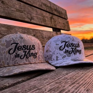 Puede incluir: Dos gorras de b&eacute;isbol con un patr&oacute;n de camuflaje en tonos beige, marr&oacute;n y blanco. Cada gorra tiene el texto "Jesus is King" bordado en letra cursiva negra en el panel frontal. La gorra de la izquierda incluye un detalle de cord&oacute;n trenzado.