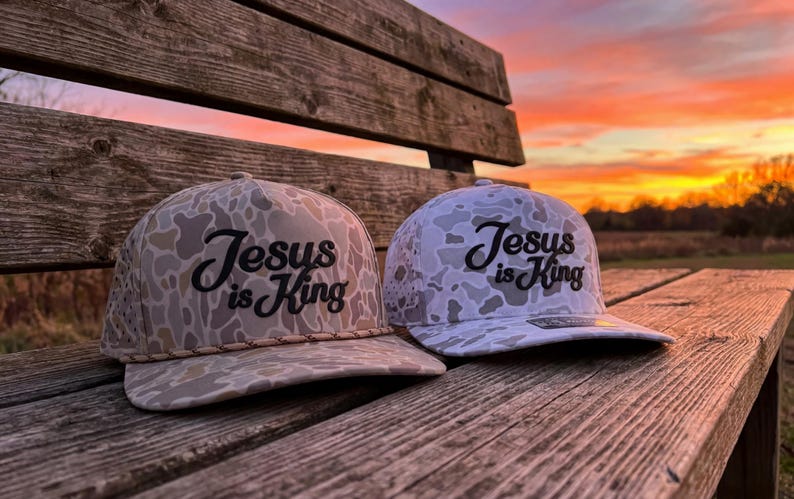 Puede incluir: Dos gorras de b&eacute;isbol con un patr&oacute;n de camuflaje en tonos beige, marr&oacute;n y blanco. Cada gorra tiene el texto "Jesus is King" bordado en letra cursiva negra en el panel frontal. La gorra de la izquierda incluye un detalle de cord&oacute;n trenzado.