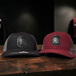 Op de afbeelding: Twee baseballpetten met mesh achterkant, één in zwart en één in bordeauxrood. Elke pet heeft een donker metalen embleem op het voorpaneel met een gestileerde letter C en een afbeelding van een vechthaan erin. De petten hebben een gestructureerde kroon.
