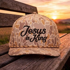 Puede incluir: Una gorra de b&eacute;isbol de camuflaje con un patr&oacute;n en tonos beige, marr&oacute;n y crema, que presenta un cord&oacute;n trenzado en la base de la corona. El panel frontal muestra el texto bordado en negro Jesus is King en una fuente cursiva estructurada.